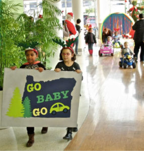 Two little girls walking in a mall holding a Go Baby Go Oregon followed by children driving switch adapted electric toy cars