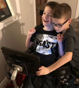 Twin boys, one in a wheelchair with a mounted communication device mounted, the other boy standing beside him touching the device.