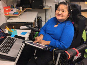 A woman is at work sitting in her wheel chair at work desk with a laptop computer, using here hand to activate an iPad communication device.